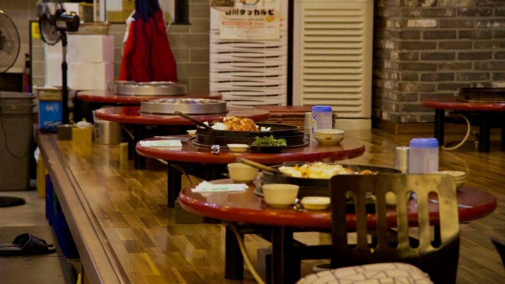 Traditional floor seating tables with round griddle pans and side dishes fill a dakgalbi restaurant.