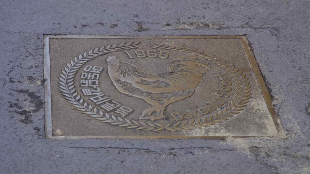 A square bronze pavement plaque labeled Dakgalbi Street with a rooster emblem and 1968 is set into the lane.
