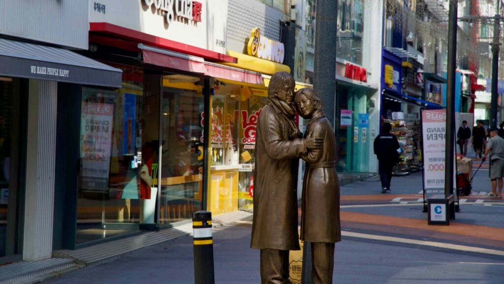 A bronze couple statue stands on the shopping street amid cafés and brand storefronts in central Chuncheon.