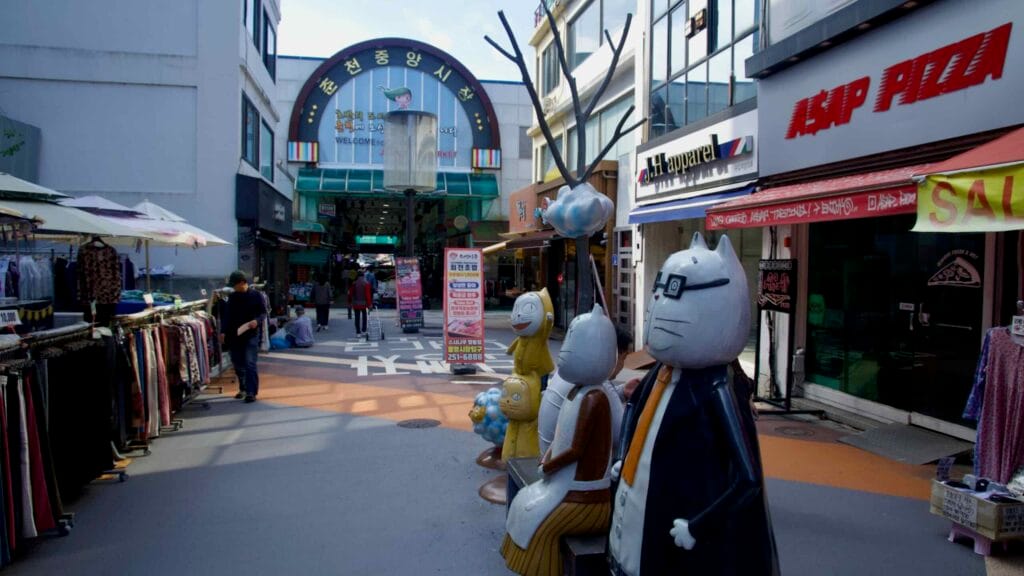 The entrance to Chuncheon Jungang Market has character statues and clothing racks along the approach.
