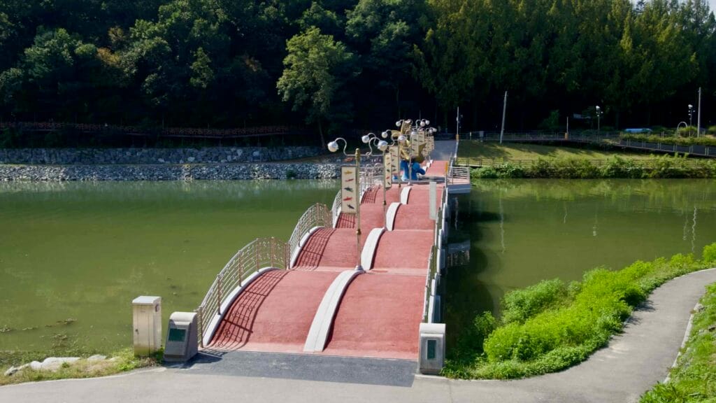 An undulating red pedestrian bridge spans Gongji Stream, with lamps and fish banners leading toward wooded banks and riverside paths.