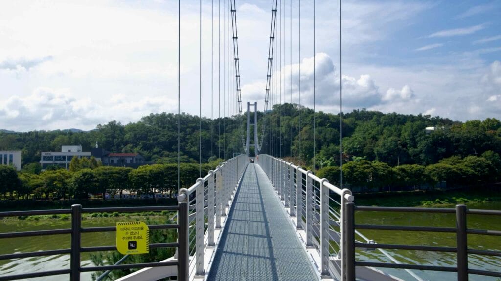 Pedestrians cross Chuncheon’s Sairo 248 suspension bridge above Uiam Lake and low forested hills.