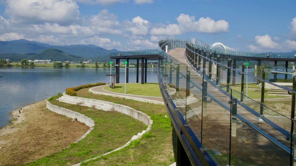 Glass-railed walkways curve over lawns at Munhwa Gwangjang Forest beside Lake Uiam.