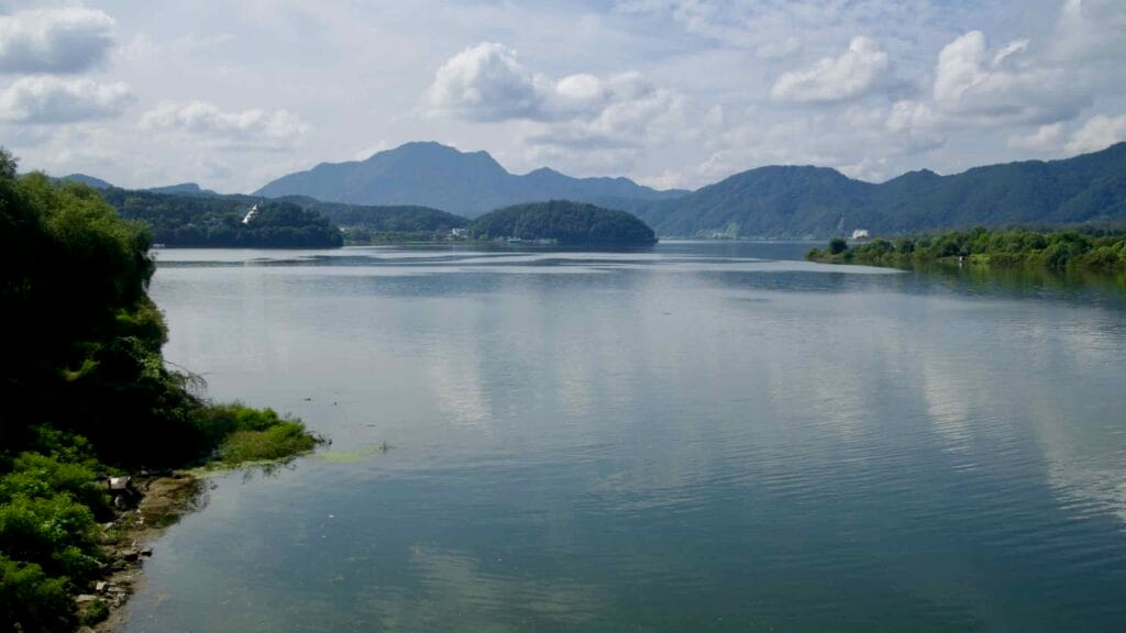 Visitors stroll through a lakeside park in Chuncheon beside trees, lawns, and the edge of the reservoir.