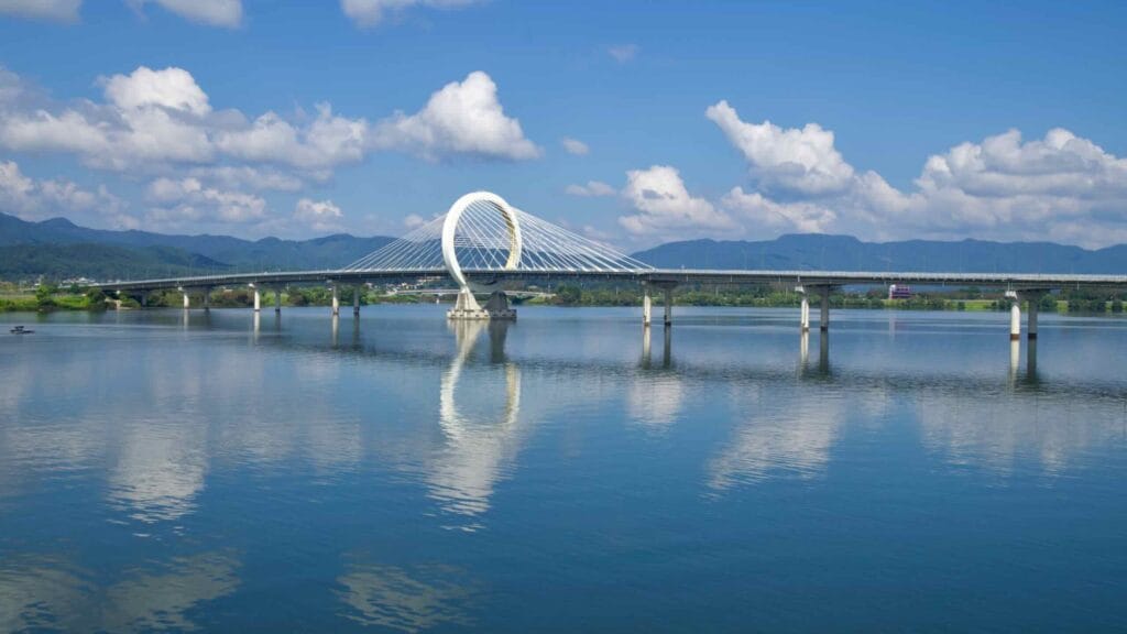 Cyclists and walkers share a lakeside park path in Chuncheon beside open lawns and water.