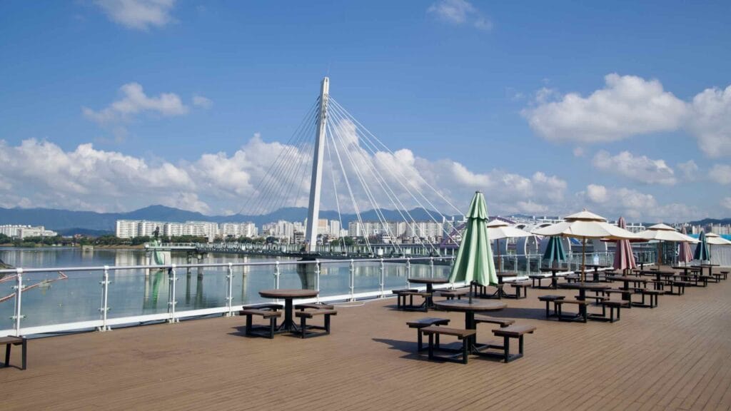 Café tables and sun umbrellas line Soyang River Skywalk’s deck, overlooking calm Uiam Lake and distant hills.
