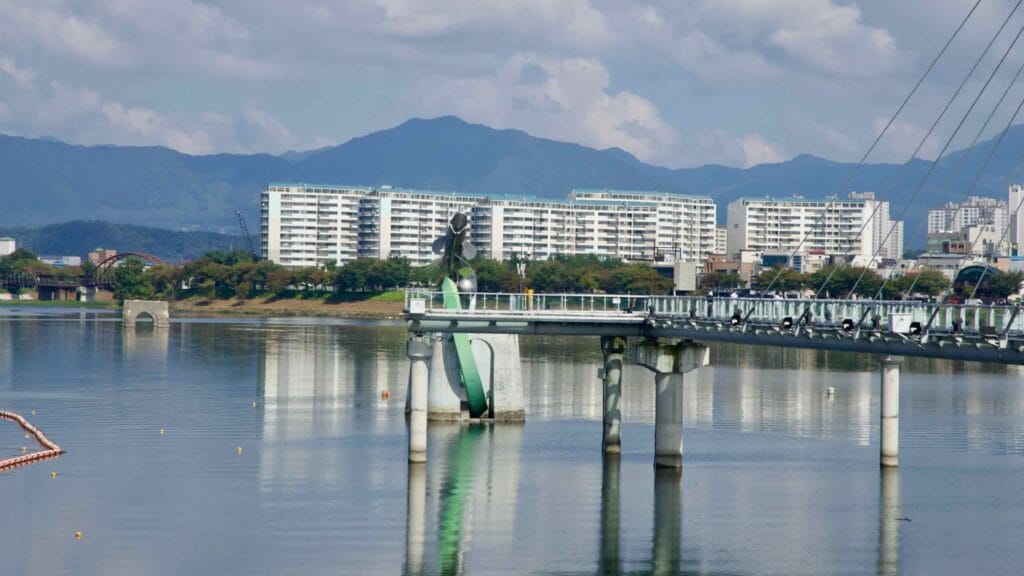 Soyang River Skywalk stretches over Uiam Lake with apartments and mountain ridges reflected in the calm water.