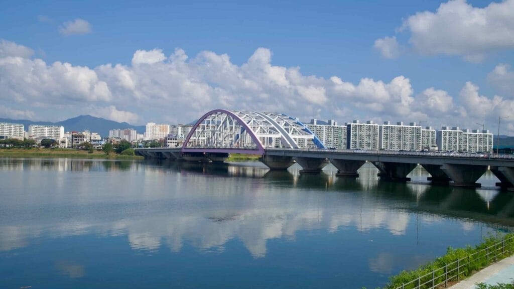 A multicolored steel arch road bridge crosses the river in Chuncheon, with apartments and city buildings along the bank.