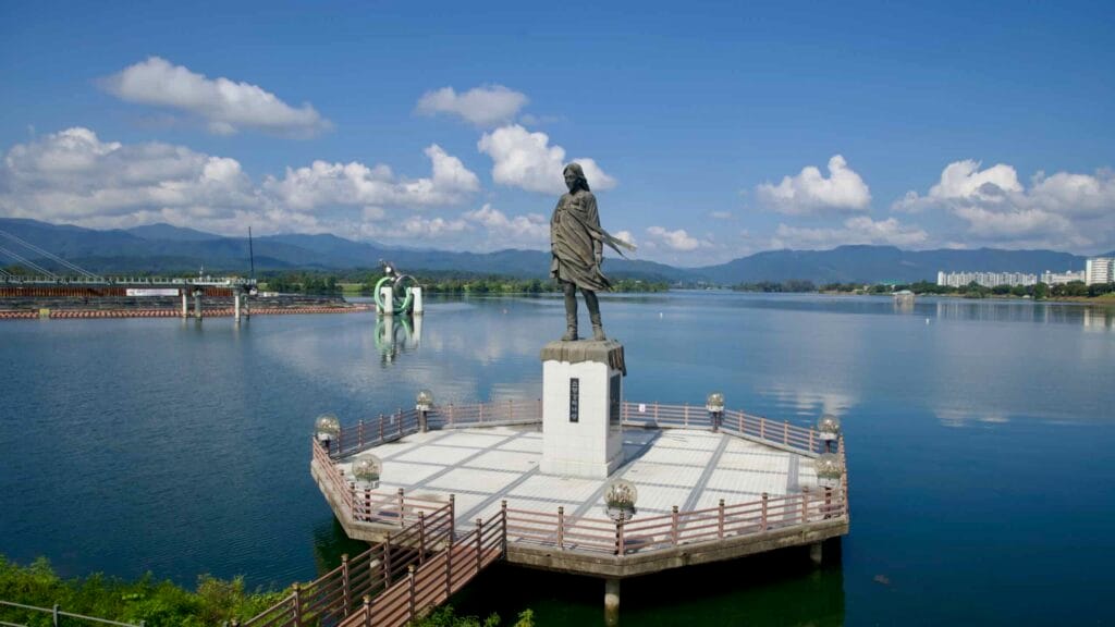 The Soyang River Maiden statue stands on an octagonal deck in Lake Uiam, with mountains and the fish sculpture beyond.