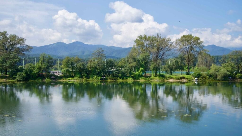 Trees and low mountains reflect across a calm river bend near Chuncheon, where anglers line the shore.