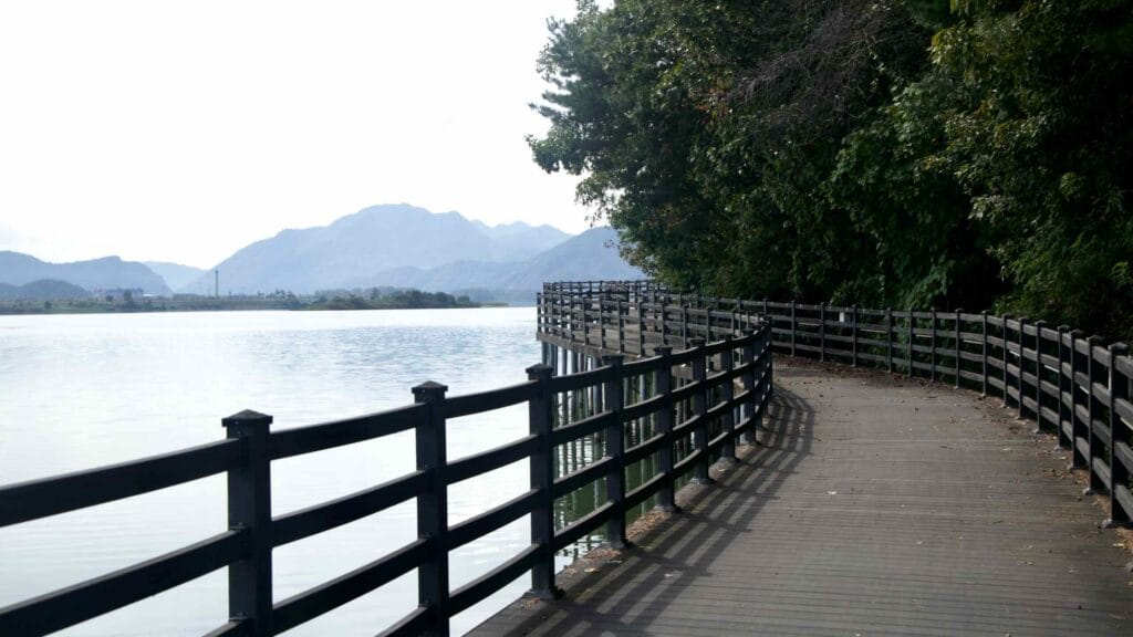 Long curving boardwalk above calm water with layered mountains ahead.