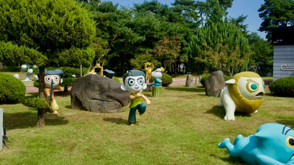 Wide view of cartoon figures around boulders and pines in the museum garden.