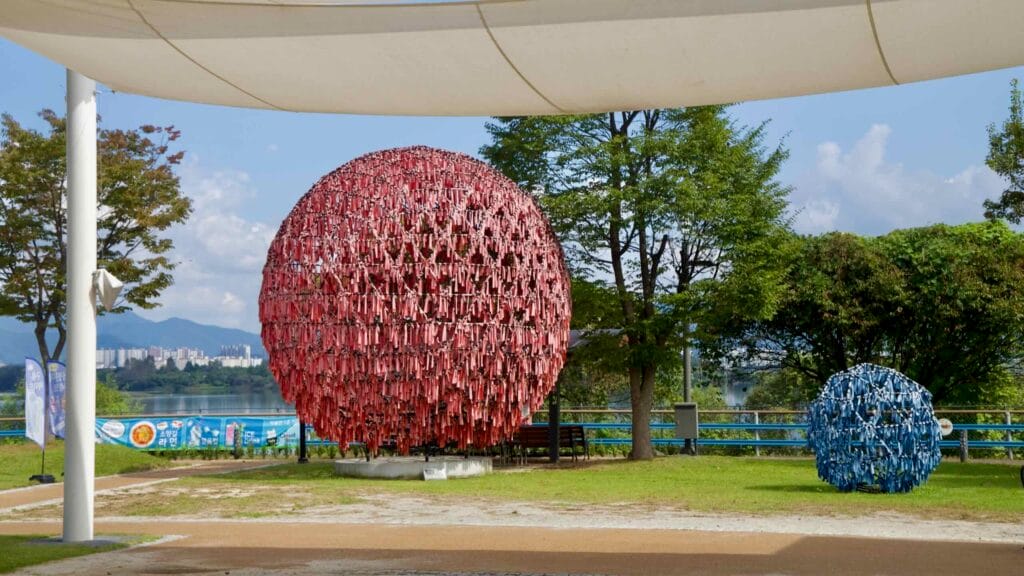 Red and blue ribbon sphere artworks under a shade canopy by the lake.