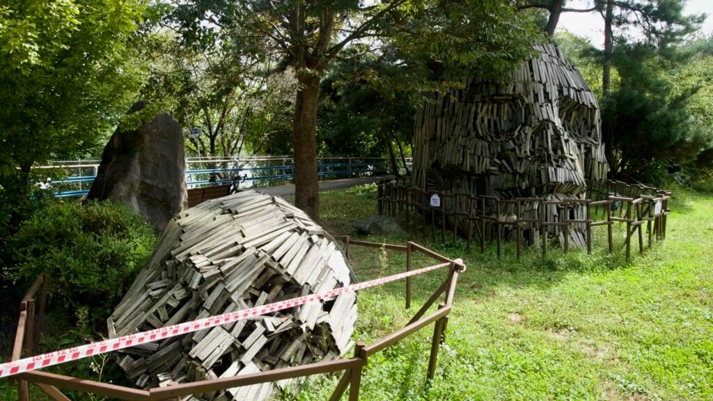 Two weathered wooden head sculptures, one upright and one fallen, sit behind fencing in the riverside grove.