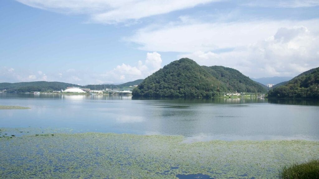 Uiam Lake widens toward Samaksan and a distant white dome, with water lilies floating in the foreground shallows.