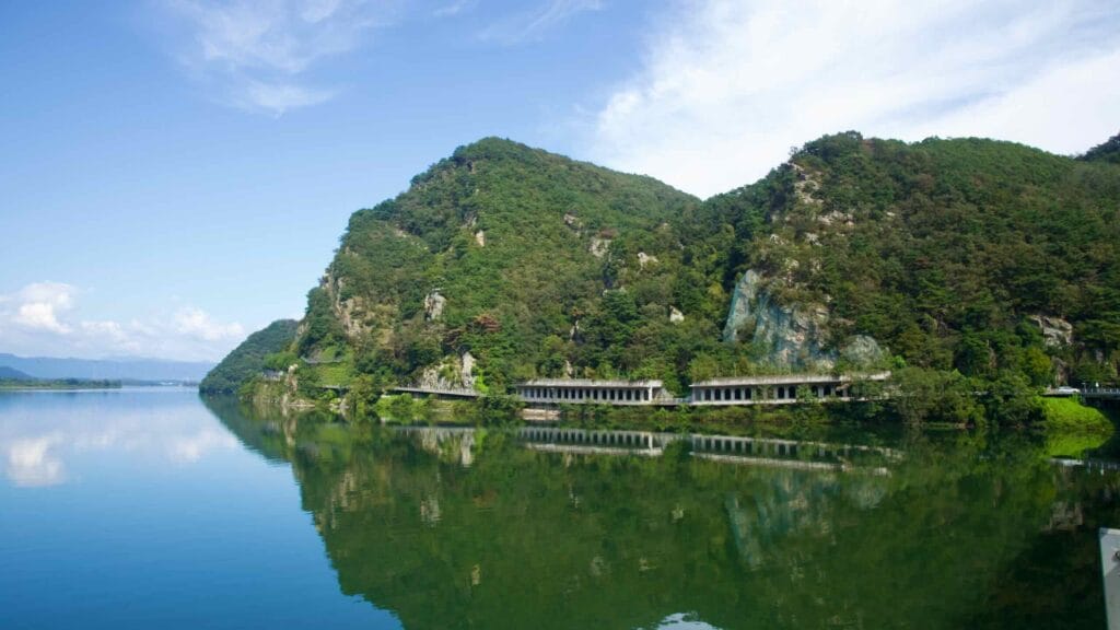 Arcaded cliffside galleries and a narrow road trace the green shoreline of Uiam Lake near Samaksan in Chuncheon, with.