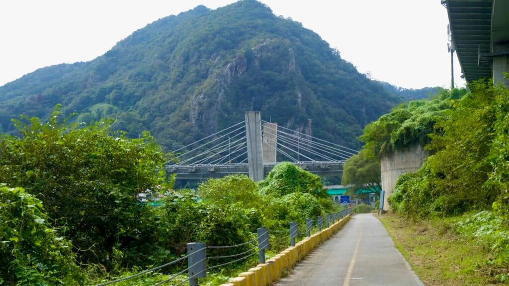 A riverside bike path curves toward Gangchon Grand Bridge beneath forested hills.