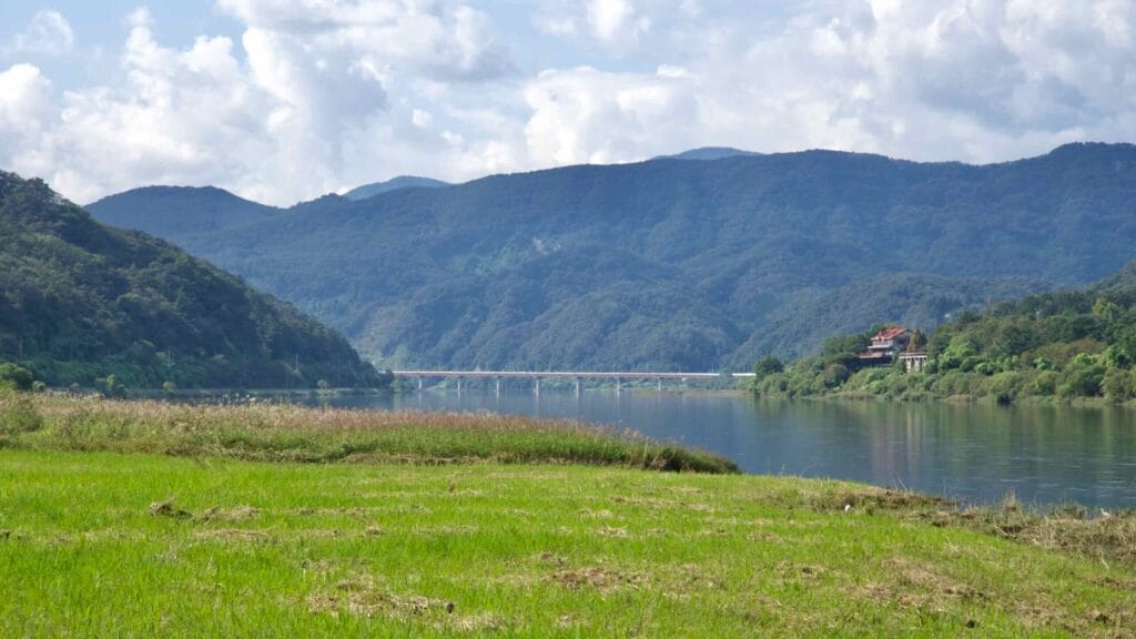 A grassy field frames the Bukhangang with a distant road bridge and layered hills.