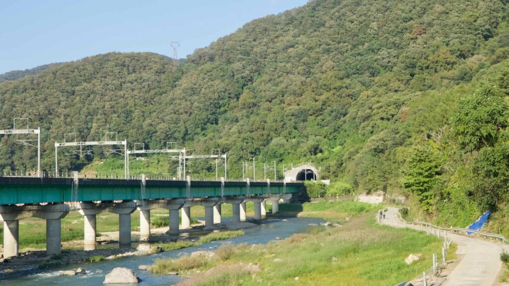 The Gyeongchun Line railway spans the Bukhan River on concrete piers toward a mountain tunnel near Cheongpyeong, with a riverside park below.