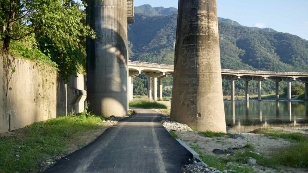 A fresh strip of asphalt threads between massive bridge piers on the riverside path below Cheongpyeong 1 Bridge, with mountains rising ahead.